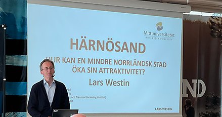 Man som står upp och talar inför publik. Bild som visas i bakgrunden där det står "Hur kan en mindre norrländsk stad öka sin attraktivitet?"