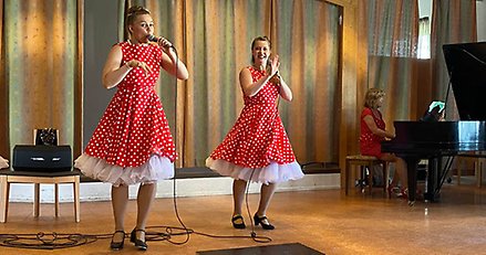 Två kvinnor i röda klänningar sjunger och dansar. I bakgrunden syns en kvinna i rött som spelar flygel.