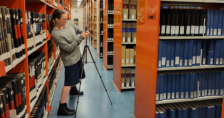 En kvinna förbereder för en filminspelning i en arkivlokal med många pärmar, genom att fästa en mobiltelefon på ett kamerastativ.