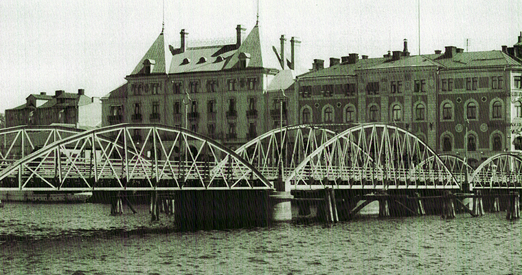 gammal svartvit bild som visar en bro med tre bågar på vardera långsida. I bakgrunden stora hus.