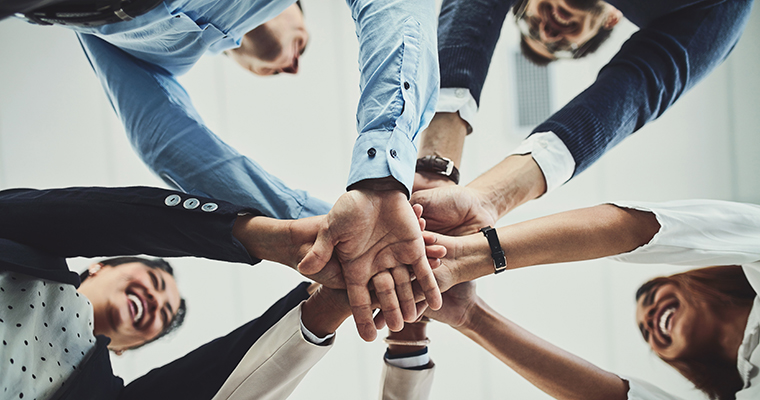 Glada människor i en ring håller in varsin hand mot cirkelns mitt som en symbol för enighet och lagarbete.
