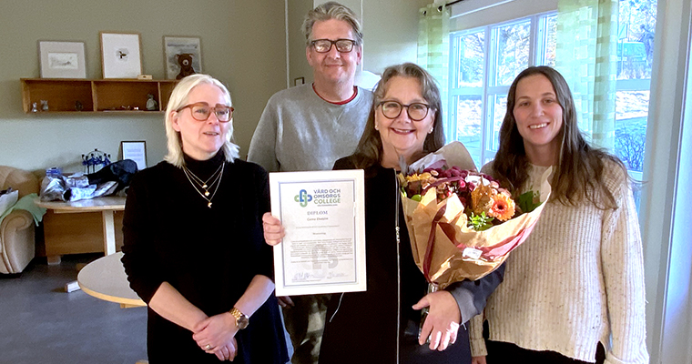 Fyra leende personer, en man och tre kvinnor. Kvinnan i mitten visar upp ett diplom och en blombukett.