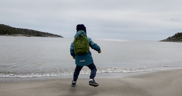ett barn med ryggsäck på ryggen ses bakifrån och hoppar på ett ben på en sandstrand med havet i bakgrunden