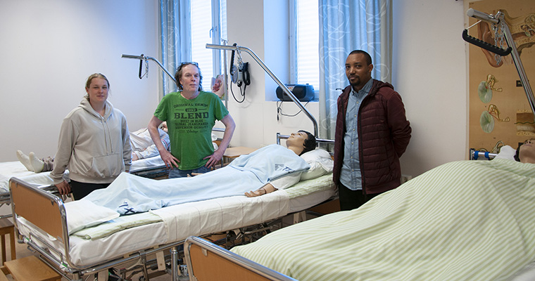 tre personer står runt en sjukhussäng med en patient i form av en fullstor docka.