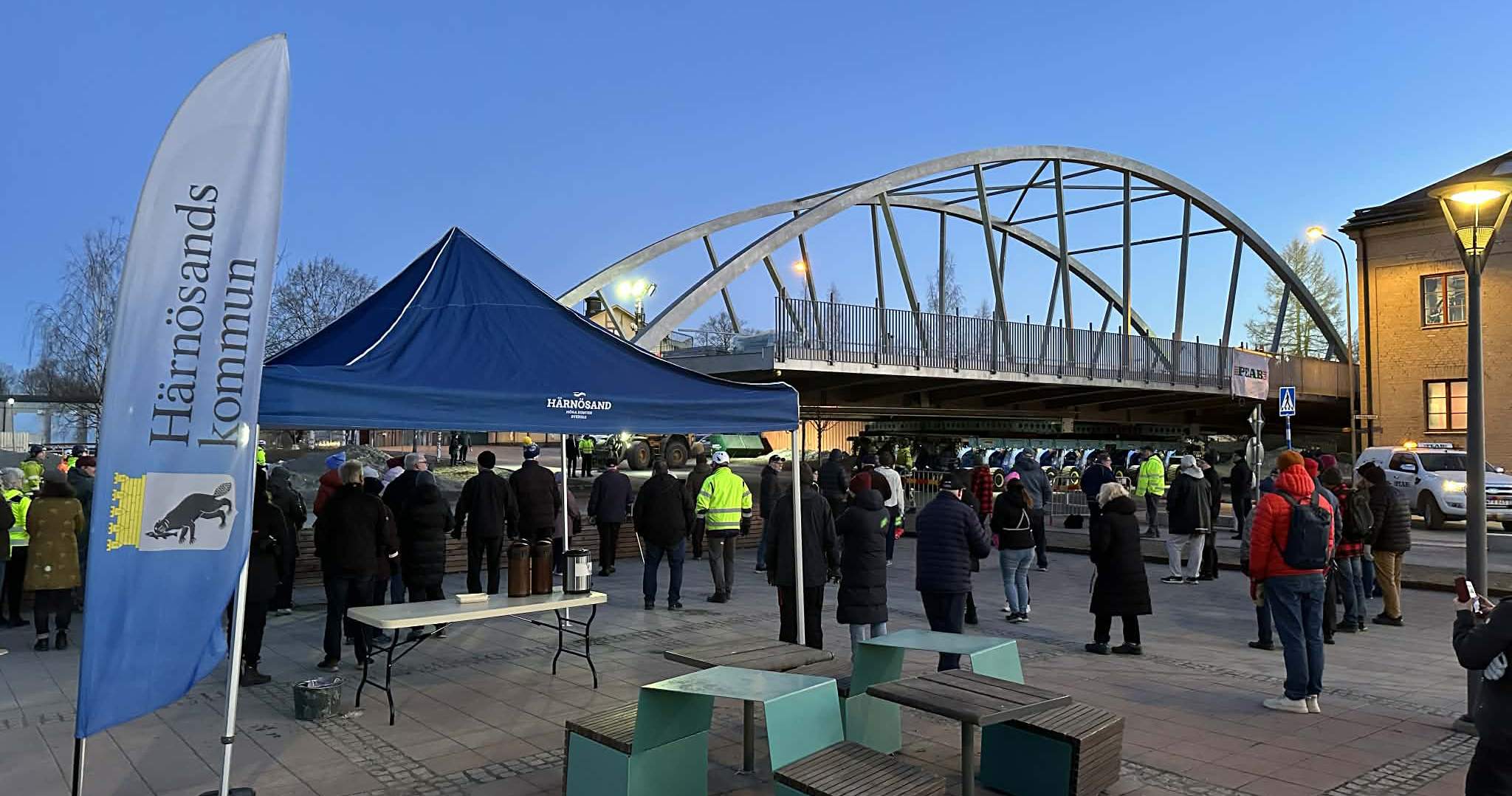 stålbro fraktas på trailer i bebyggelse, människor står bredvid och tittar på