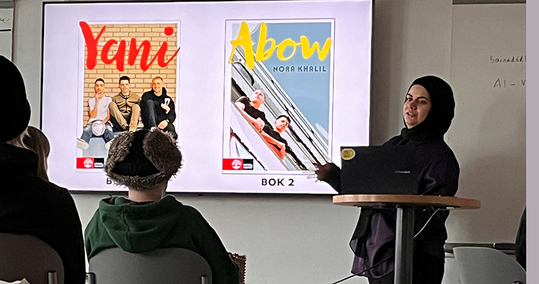 En ung kvinna visar två bokomslag på en bildskärm och berättar för elever som sitter med ryggen mot kameran.