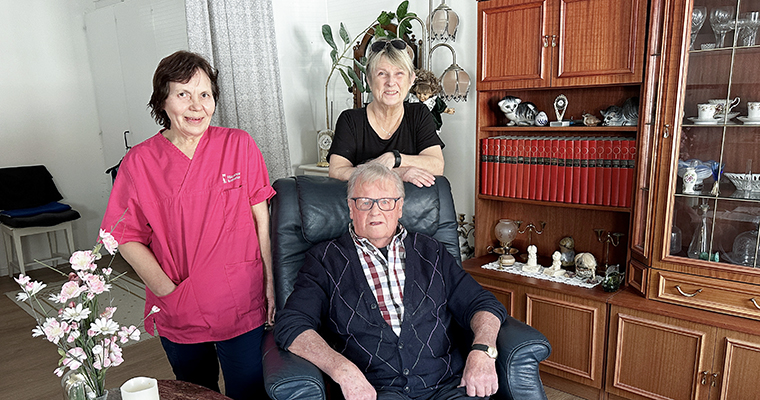 en äldre man sitter i en fåtölj i ett vardagsrum. bakom fåtöljen står två kvinnor