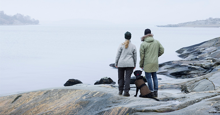 Två vuxna och en hund som står på strandområde och tittar ut mot vattnet.