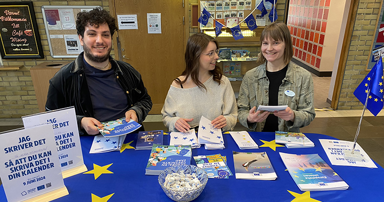 En man och två kvinnor står vid ett bord med en EU-flagga som duk. På bordet finns många informationsbroschyrer.