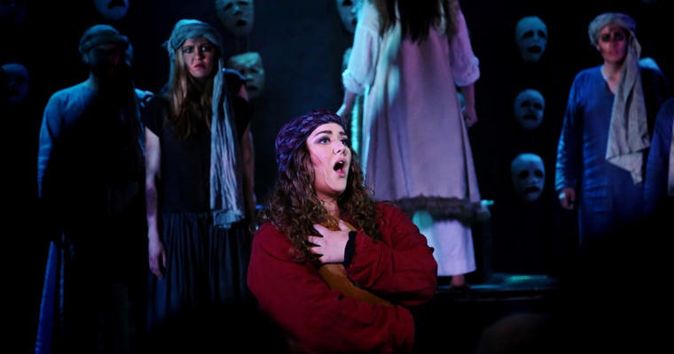 Teaterföreställning. Kvinna i rött sjunger på scen med ansikten i bakgrunden.