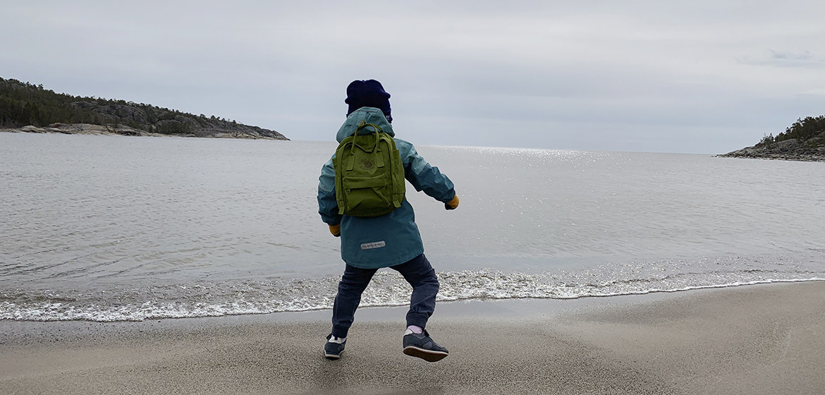 ett barn med ryggsäck sedd bakifrån gör ett litet hopp på en sandstrand med havet i bakgrunden