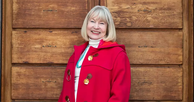 En leende äldre kvinna i röd kappa står vid en träpanel,