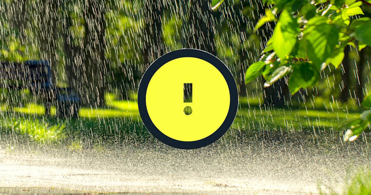 Kraftigt regn på vägbana med grön skog i fonden. En vädervarningssymbol med ett utropstecken på gul botten.