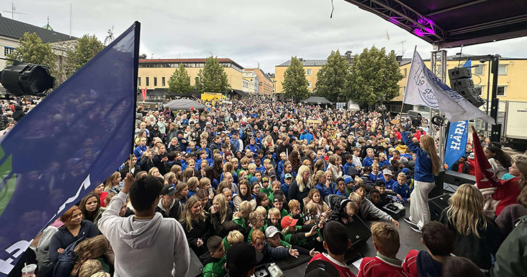 Stor folksamling framför en scen. På scenen står två personer med föreningsflaggor i händerna.