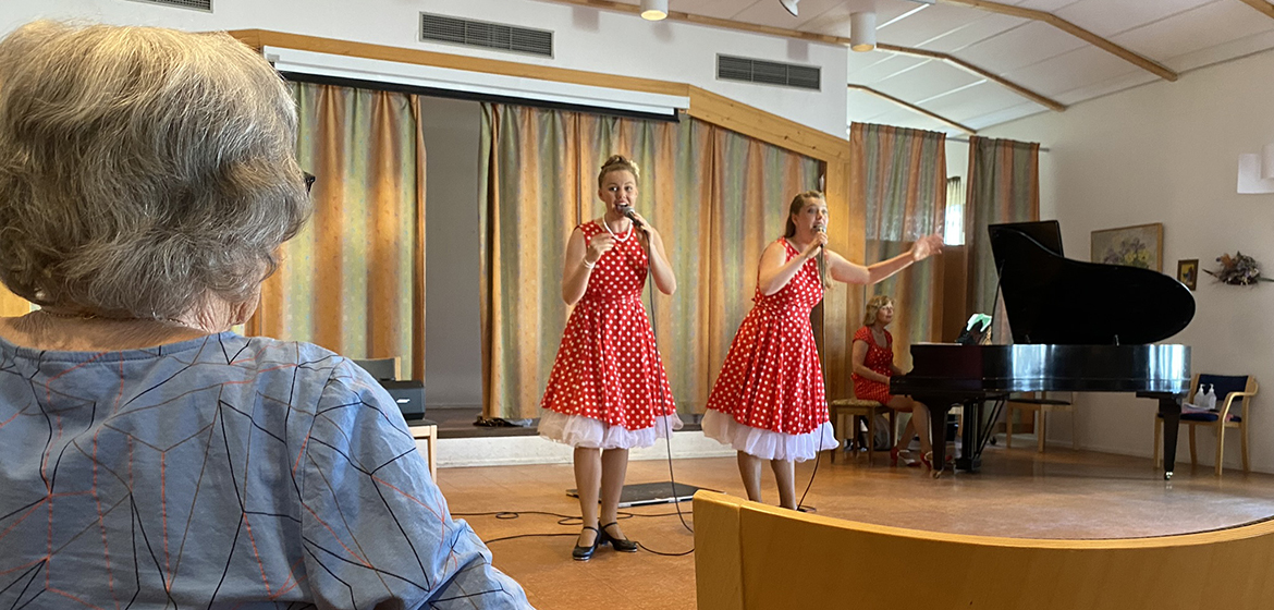 Två tjejer i röd klänningar sjunger och showar. I förgrunden syns en oidentifierbar äldre kvinna.