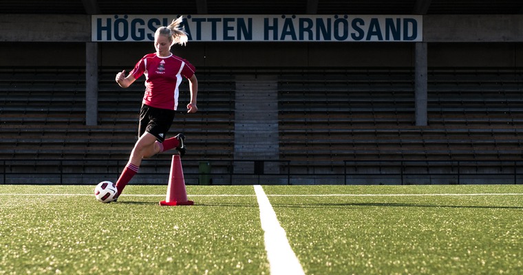 ung kvinna springer med en fotboll på en fotbollsplan. I bakgrunden en skylt där det står Högslätten.