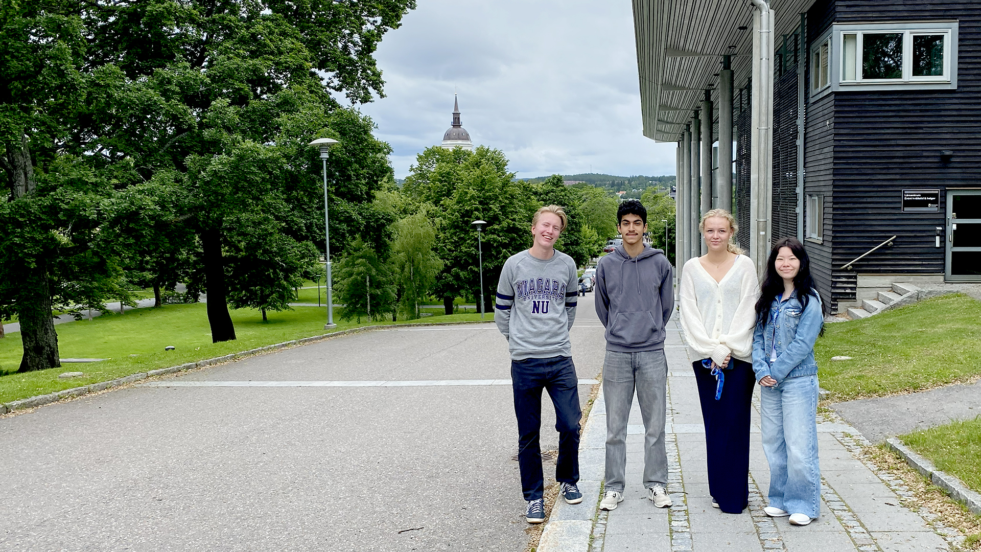 Fyra leende ungdomar vid Sambiblioteket i Härnösand, med stadsparken och domkyrkan i fonden. 