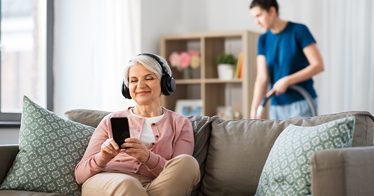 Äldre kvinna i soffa lyssnar på musik i lurar medan en ung kvinna dammsuger i bakgrunden.