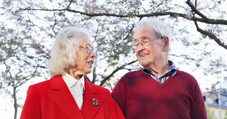 ett äldre par, en kvinna och en man, ser på varandra och ler. I bakgrunden finns ett träd