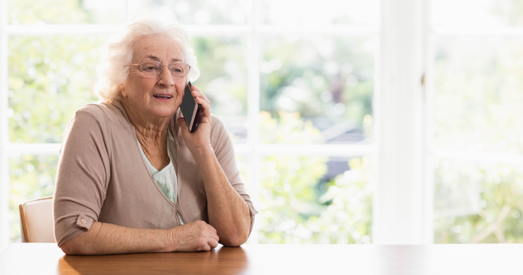 Äldre kvinna som sitter vid ett bord och pratar i en mobiltelefon.