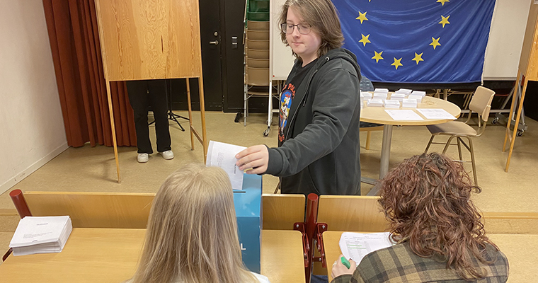 En högstadiekille lägger sin röst i en valurna medan två oidentifierbara valförrättare tittar på. I bakgrunden en EU-flagga.