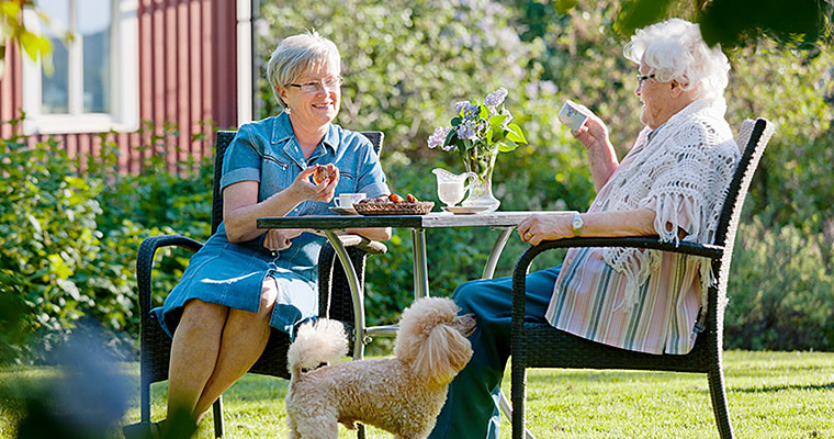 Två äldre kvinnor som i fikar i trädgården, hunden gör dem sällskap.