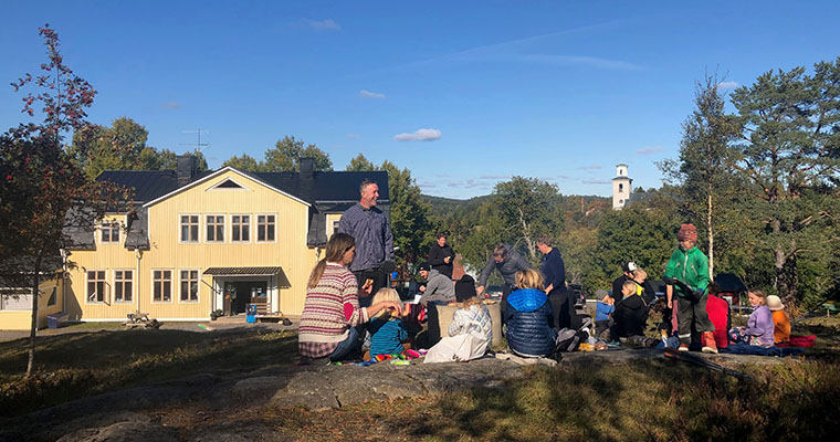 Barn och vuxna sitter tillsammans på en stenklippa, Häggdångers byskola och Häggdångers kyrkas kyrktorn i fjärran.