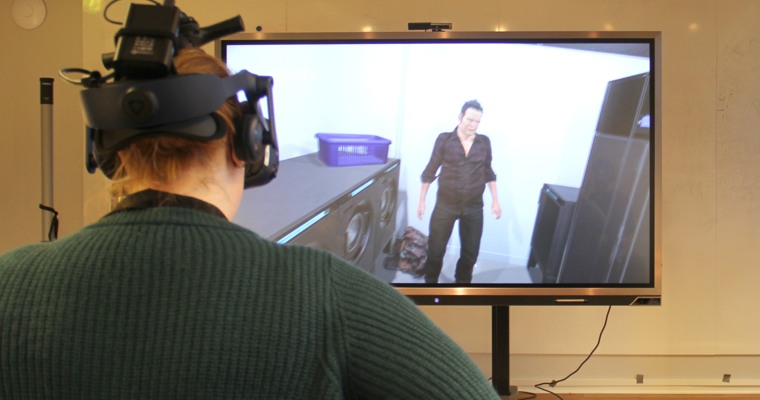 En kvinna med vr-headset på huvudet står vänd mot en tv-skärm som visar en datoranimerad bild på en man i ett rum.