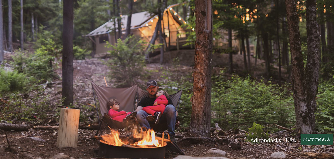 en man och två barn sitter i campingstolar vid en lägereld i skogen. I bakgrunden syns en stuga.