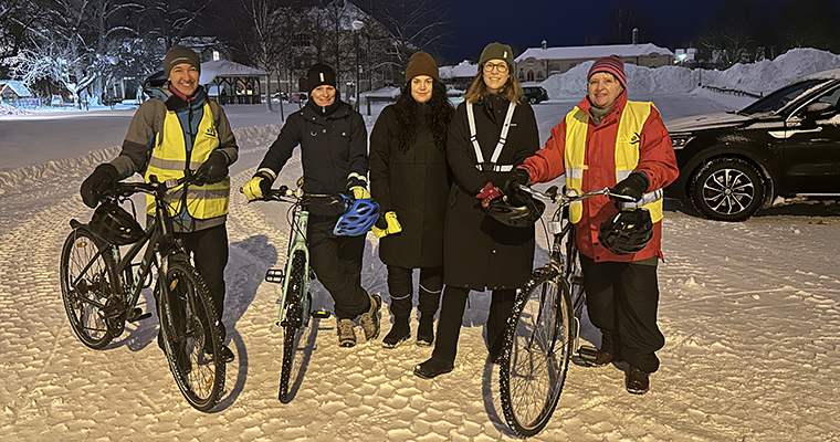 fem personer står med reflexvästar och cyklar på en snöig parkering