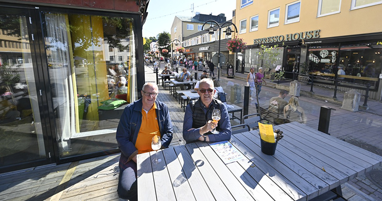 två män sitter vis ett bord på en uteservering i stadsmiljö