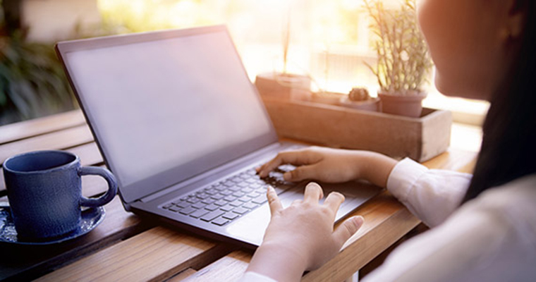 Genrebild som visar en oidentifierbar person som sitter och arbetar på sin laptop vid ett fönster.