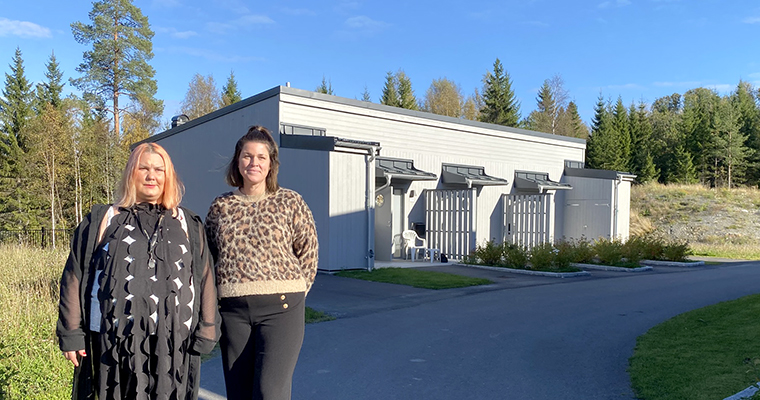 Två kvinnor står utomhus framför en låg byggnad med grå träpanel.
