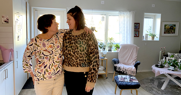 Två kvinnor som står i ett vardagsrum. De tittar på varandra och ler.