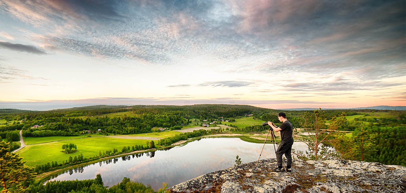 en person står med ett kamerastativ på ett berg med vid utsikt över en sjö, ängar och skog