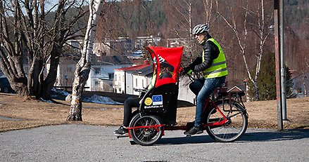 en person i gul väst cyklar på en stor cykel med en soffa fram. I soffan sitter en annan person med gul väst