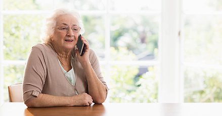 Äldre kvinna som sitter vid ett bord och pratar i en mobiltelefon.