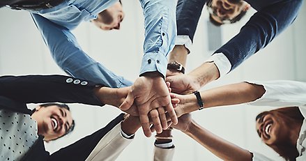 Glada människor i en ring håller in varsin hand mot cirkelns mitt som en symbol för enighet och lagarbete.