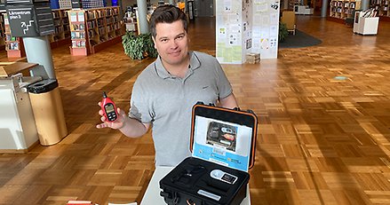 En man står i en biblioteksmiljö och visar upp en väska med mätinstrument.