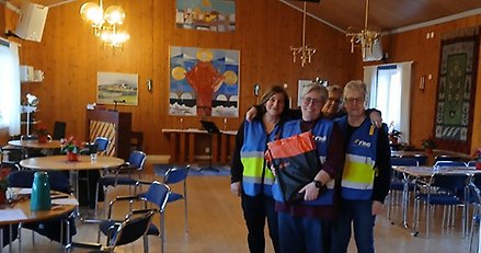 Tre leende kvinnor i reflexvästar står inomhus.