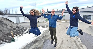 Tre mycket glada personer, en man och två kvinnor hoppar och sträcker armarna i luften. De står utomhus framför en låg gråmålad byggnad.