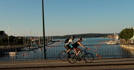 Sommar och två cyklister som cyklar över en bro. Vatten och båtar nedanför.