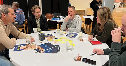 Fem personer sitter och samtalar kring ett runt bord. Papper på bordet.