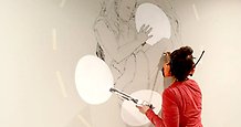 En kvinna skapar ett konstverk genom att måla på en vägg. Man ser bara konstnärens rygg.