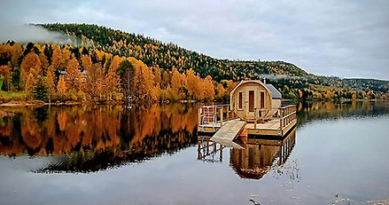 Cylinderformad bastuflotte med trädäck flyter på en spegelblank sjö med höstgula träd i bakgrunden.
