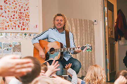 Kille sitter på en pall och spelar gitarr. Ryggar på barn i förgrunden.