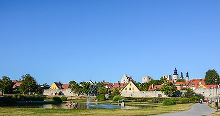 Miljöbild från Almedalen på Gotland. Damm med fontänstråle, gröna gräsmattor och gamla stenhus i bakgrunden.