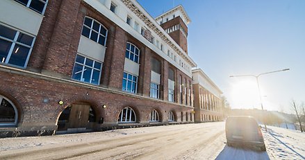 en stor röd tegelbyggnad som ligger längs en gata. På gatan står en bil parkerad och vid horisonten lyser en sol.