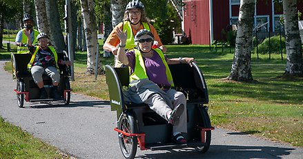 Två cyklar med stora sitsar fram. På varje cykel sitter en äldre person på sitsen som passagerare och ytterligare en person som cyklar.