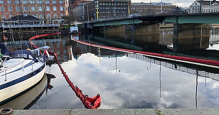 ett sund där det ligger flera långa, orange länsar tvärs över. I förgrunden en kaj och del av en båt. På andra sidan sundet hus 